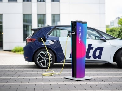 Ein Elektroauto mit blau-weißer Lackierung ist an einer modernen Ladesäule mit buntem Farbverlauf angeschlossen. Das gelbe Ladekabel verbindet das Fahrzeug mit der Station, die den Schriftzug 'CHARGE MAKER' trägt. Im Hintergrund ist ein modernes Bürogebäude mit großen Fenstern zu sehen.